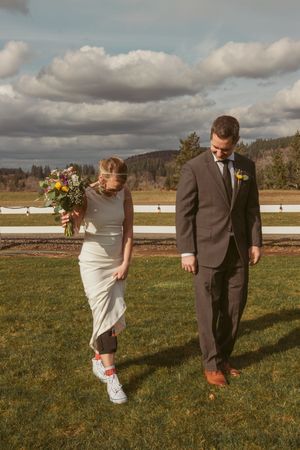 Couple walking together on grass field under dramatic cloudy sky, bride wearing white dress and sneakers.