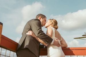 A couple shares a romantic kiss on a rooftop balcony under a cloudy sky at sunset.