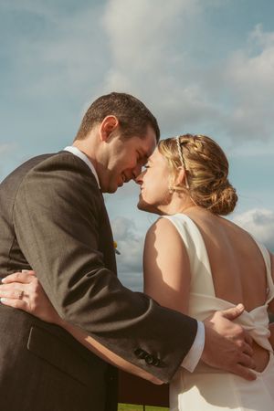 A romantic outdoor wedding photo of a couple embracing against a cloudy blue sky background.