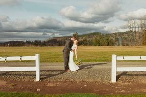 A couple shares a romantic moment between white fences at a rustic countryside venue under cloudy skies.