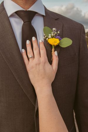 A hand wearing a wedding ring touches a brown suit lapel adorned with a yellow and purple floral boutonniere.
