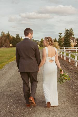 Couple walking hand in hand down a white-fenced country road at sunset after their wedding ceremony.