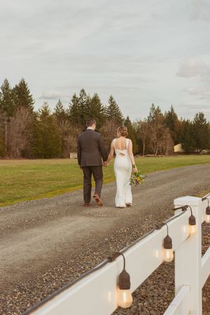 Newlyweds stroll down a country path with white fencing adorned with Edison bulbs at sunset.