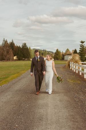 Wedding couple walking along a gravel path with evergreen trees and white fence in rural scenery.