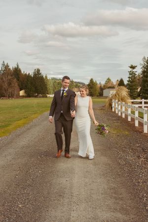 Newlyweds strolling along a gravel path lined with white fence posts under cloudy skies.