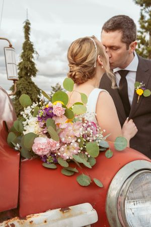 Wedding couple embraces near vintage red truck with beautiful floral bouquet of pink and white flowers.