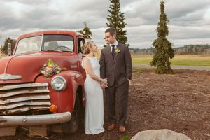 Newlyweds pose beside classic red pickup truck in rustic outdoor setting with evergreen trees.