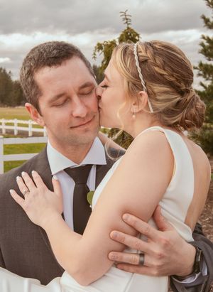A romantic wedding moment captured outdoors with gentle embrace and kiss against a cloudy sky and white fence backdrop.