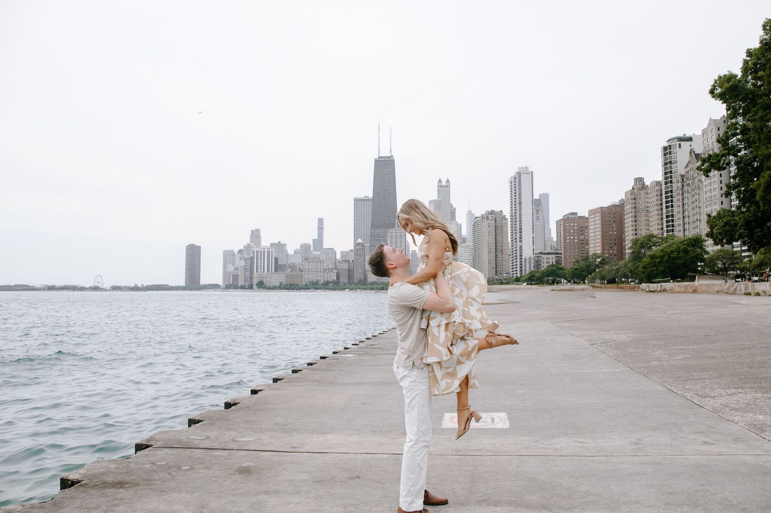 A couple in light colored outfits embrace on North Avenue Beach with the Chicago skyline in the background.