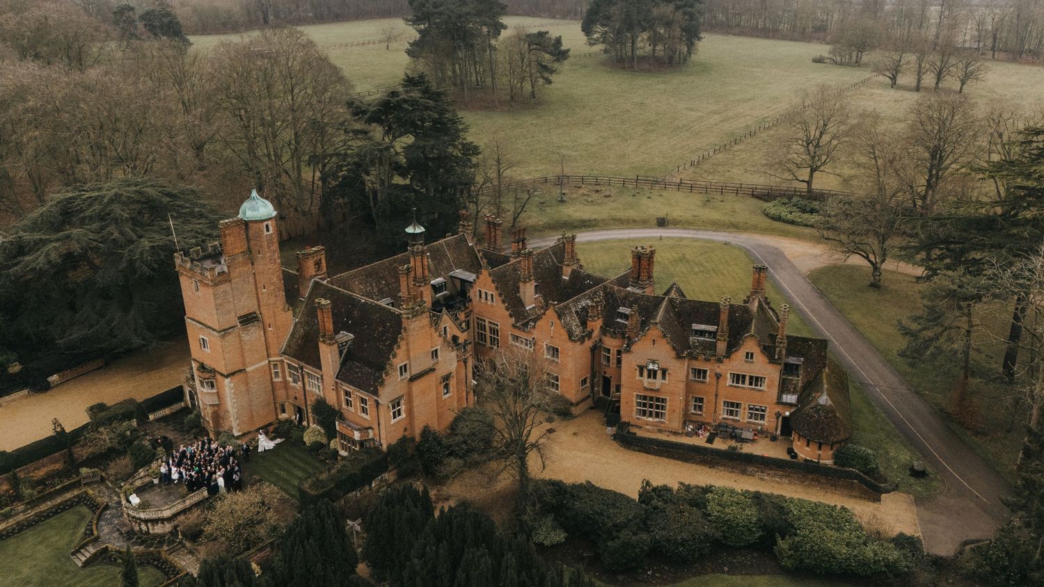 Aerial view of a grand Victorian Gothic red brick manor house with turrets and chimneys surrounded by scenic grounds.