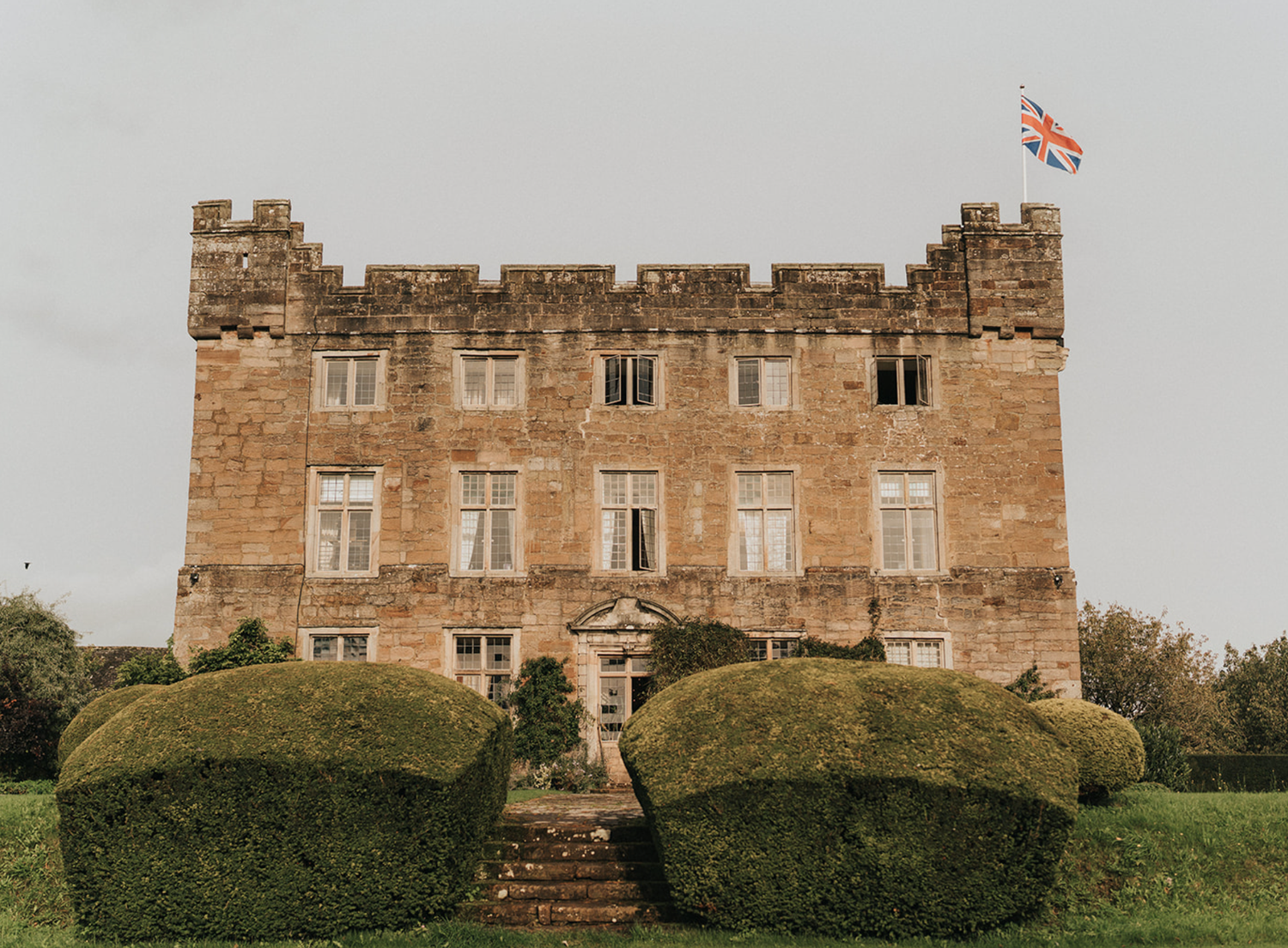 Historic stone castle with ornate facade, manicured hedges and stone steps leading to entrance, flying a red flag atop.
