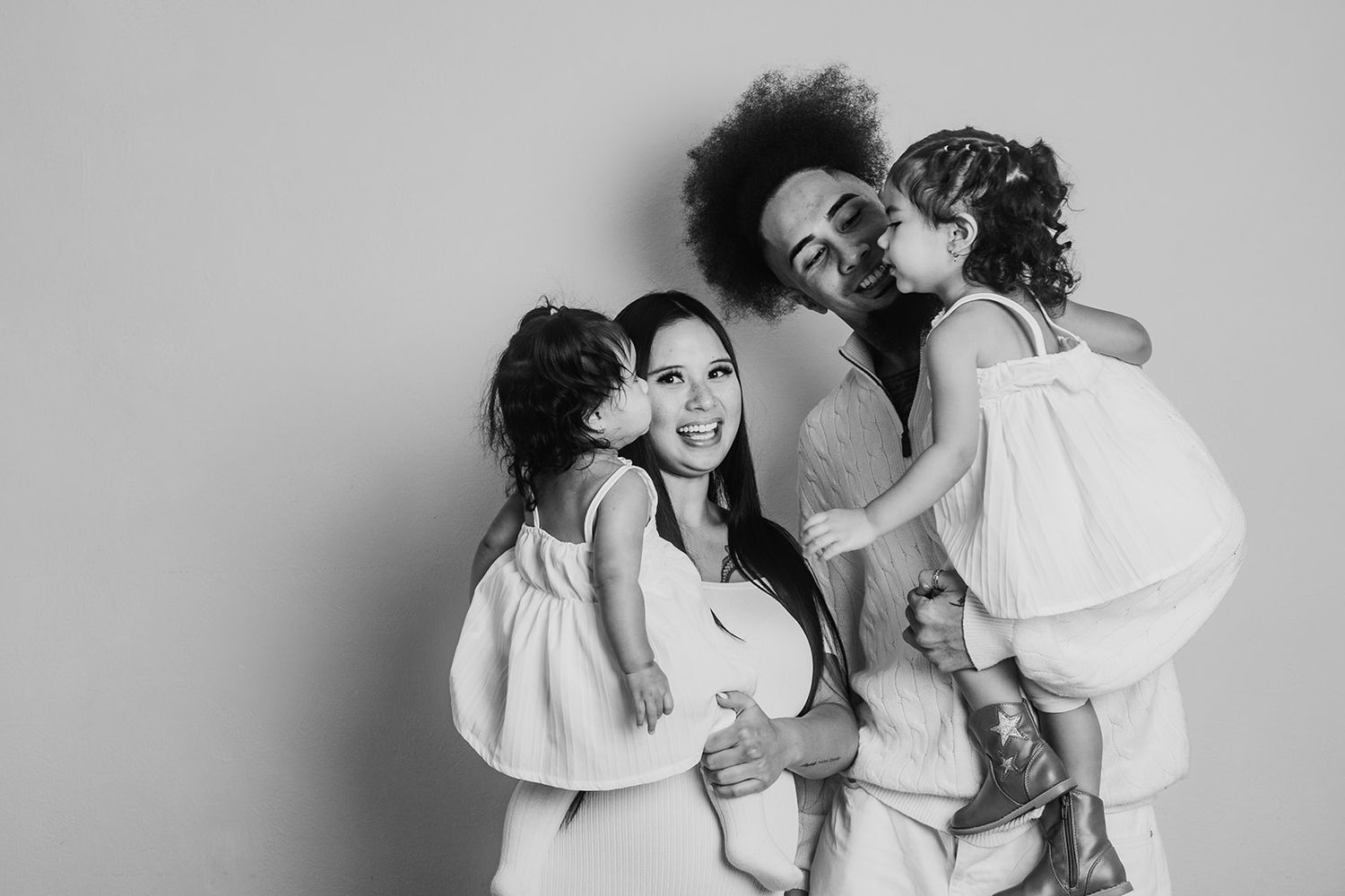 Black and white family photograph showing affectionate group embrace wearing matching white outfits against plain wall.