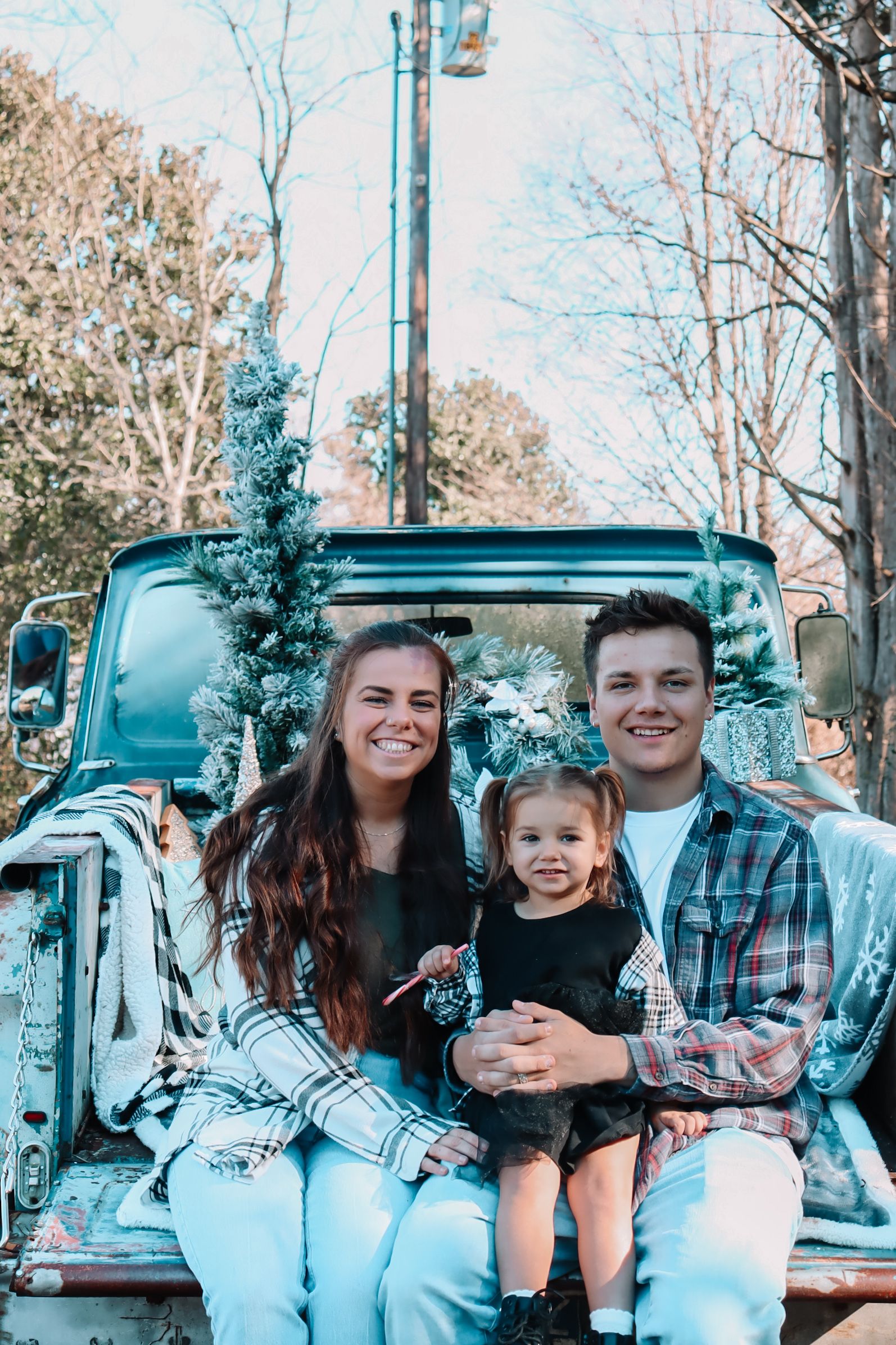 A cheerful group sits on the tailgate of a vintage blue truck with holiday decorations in a winter outdoor setting.