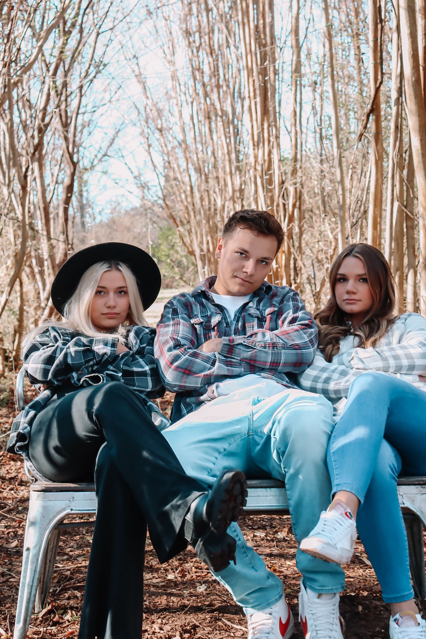 Three young people sitting together on outdoor furniture in a wooded area wearing casual outfits.