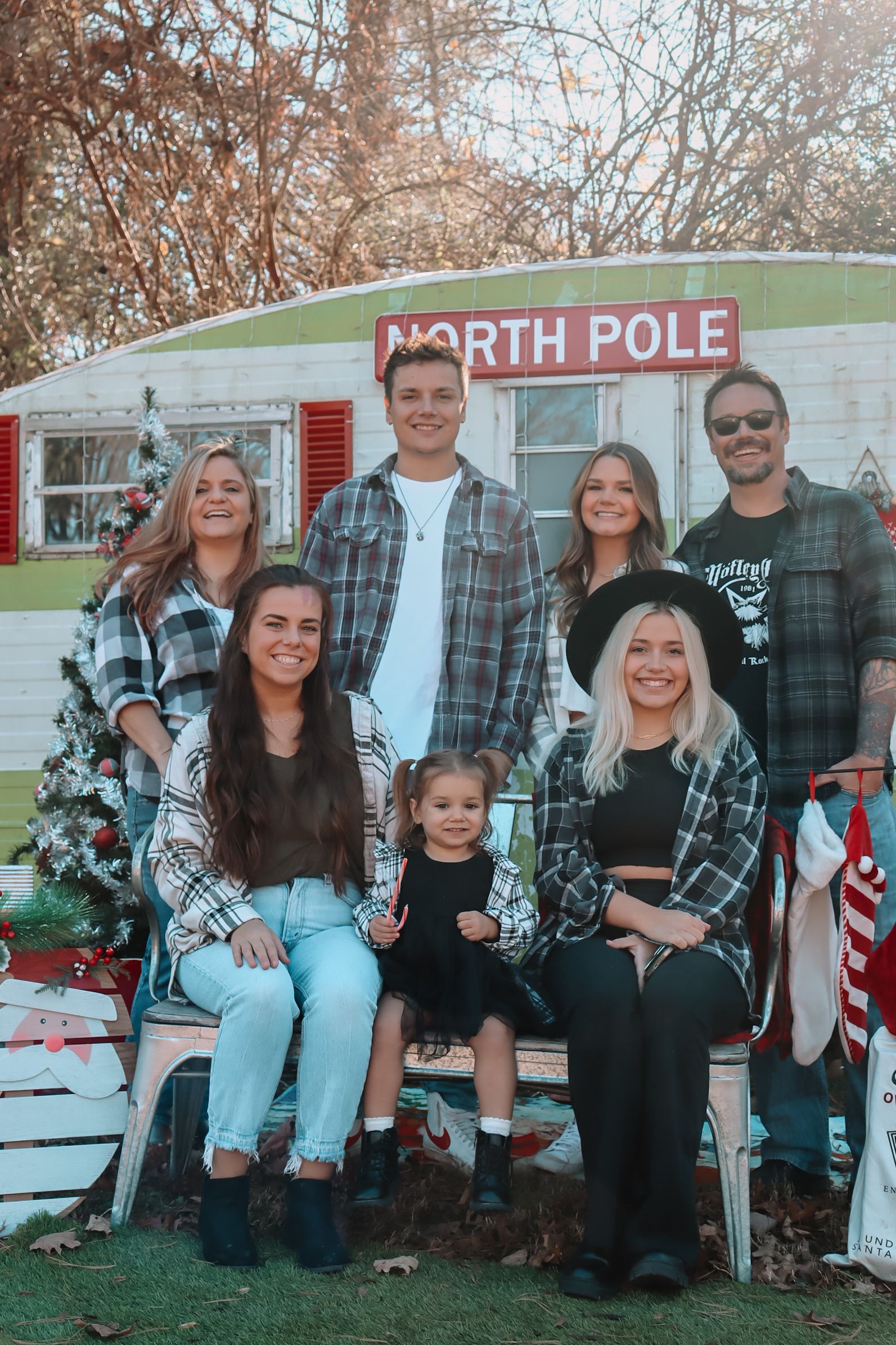 Group wearing coordinated plaid flannel shirts poses in front of rustic North Pole display with Christmas decorations.
