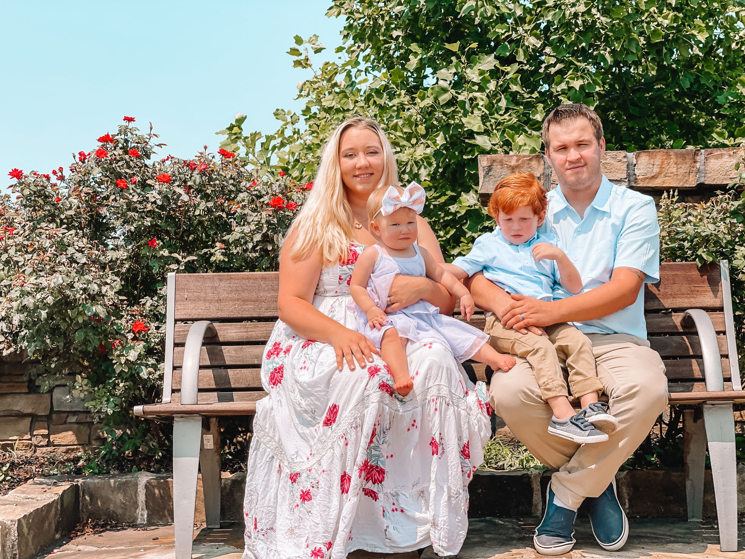 A family sits together on a wooden bench in a garden with blooming rose bushes on a sunny summer day.
