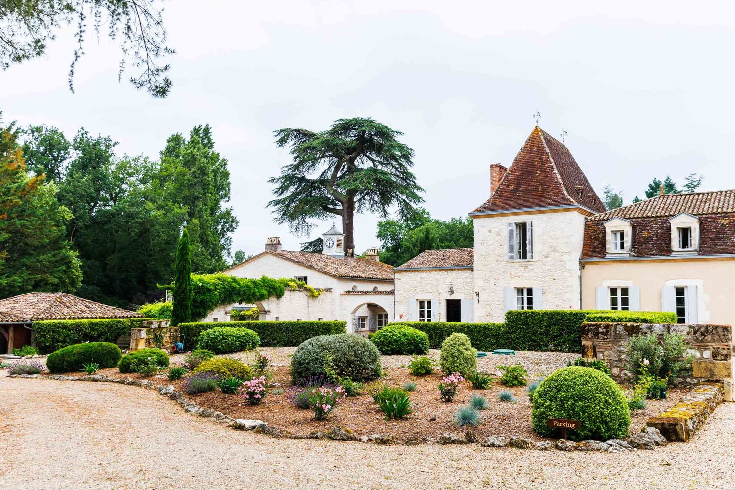 A charming French chateau with white stone walls and dormers sits among manicured gardens and gravel paths in the countryside.