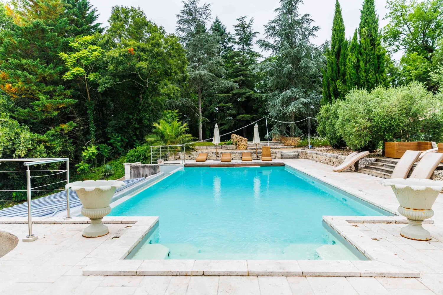 Luxurious outdoor swimming pool with stone patio, lounge chairs and evergreen trees in landscaped backyard setting.