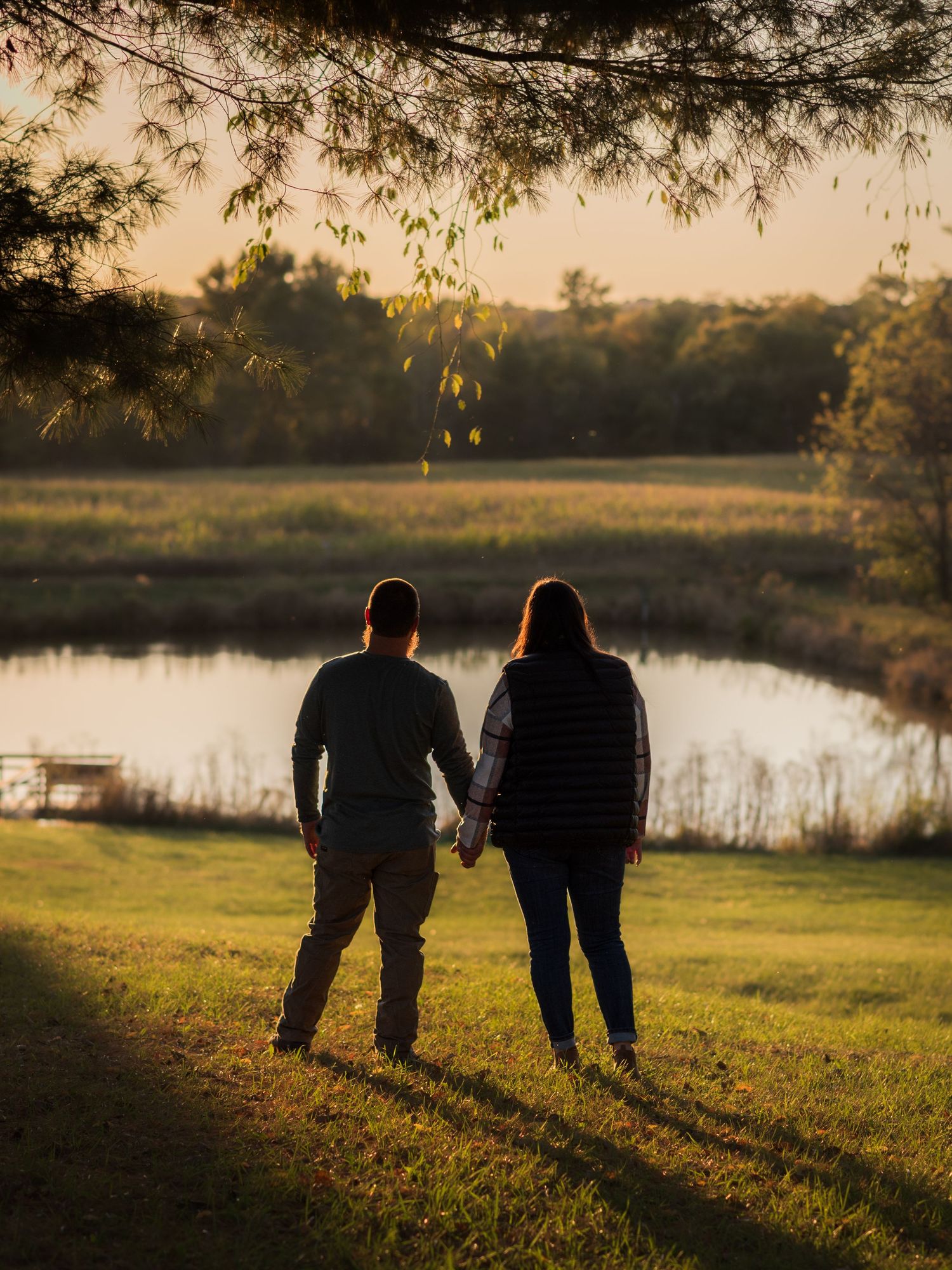 A couple sits together on a grassy hill overlooking a serene lake at golden hour.