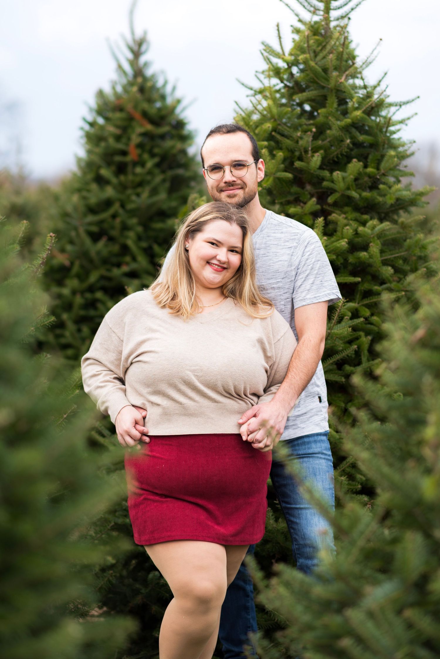 Playful couple poses together among evergreen trees at a Christmas tree farm.
