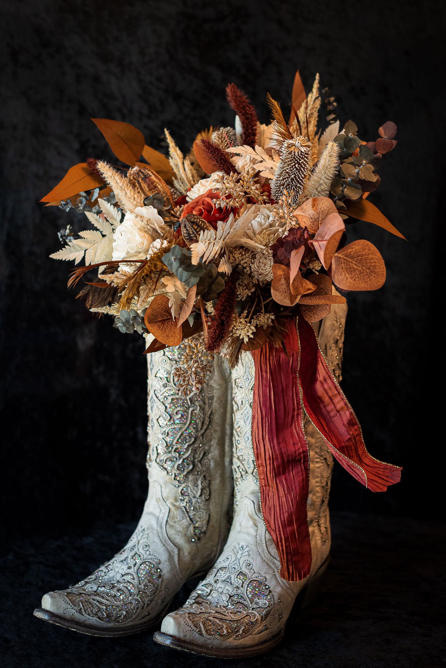 Decorative white cowboy boots adorned with autumn floral arrangements and red ribbons against a dark background.