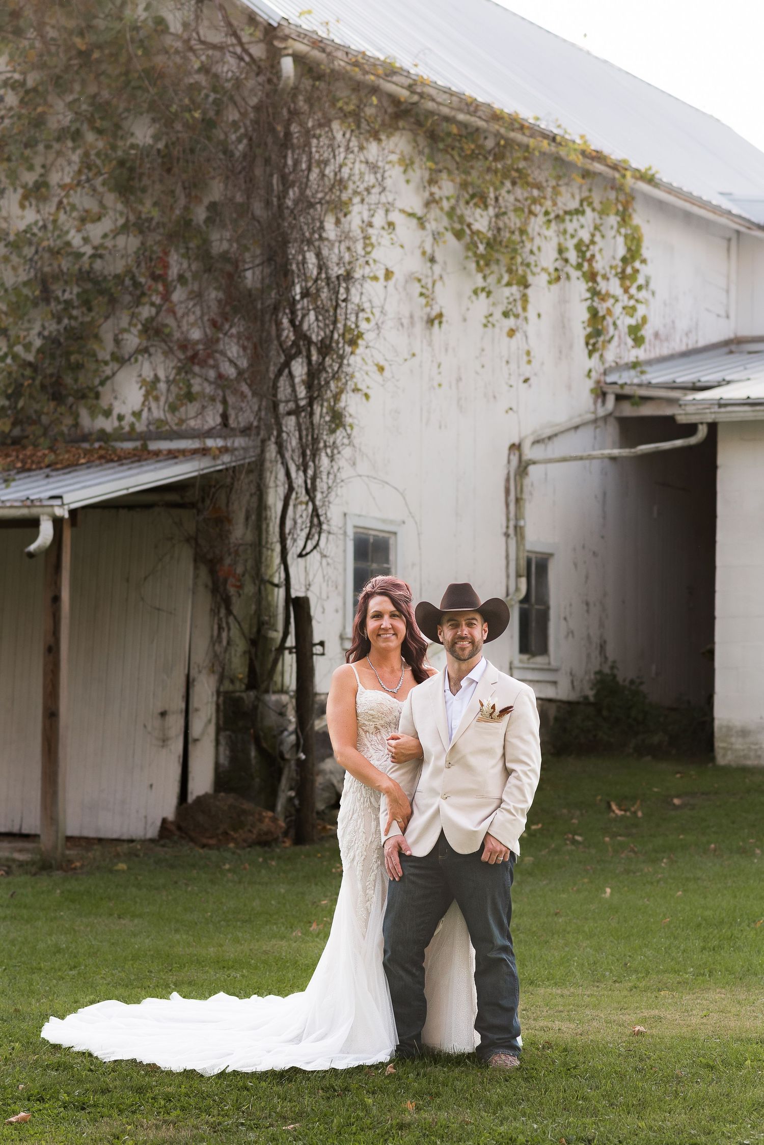A couple poses against a white barn wall with climbing vines.