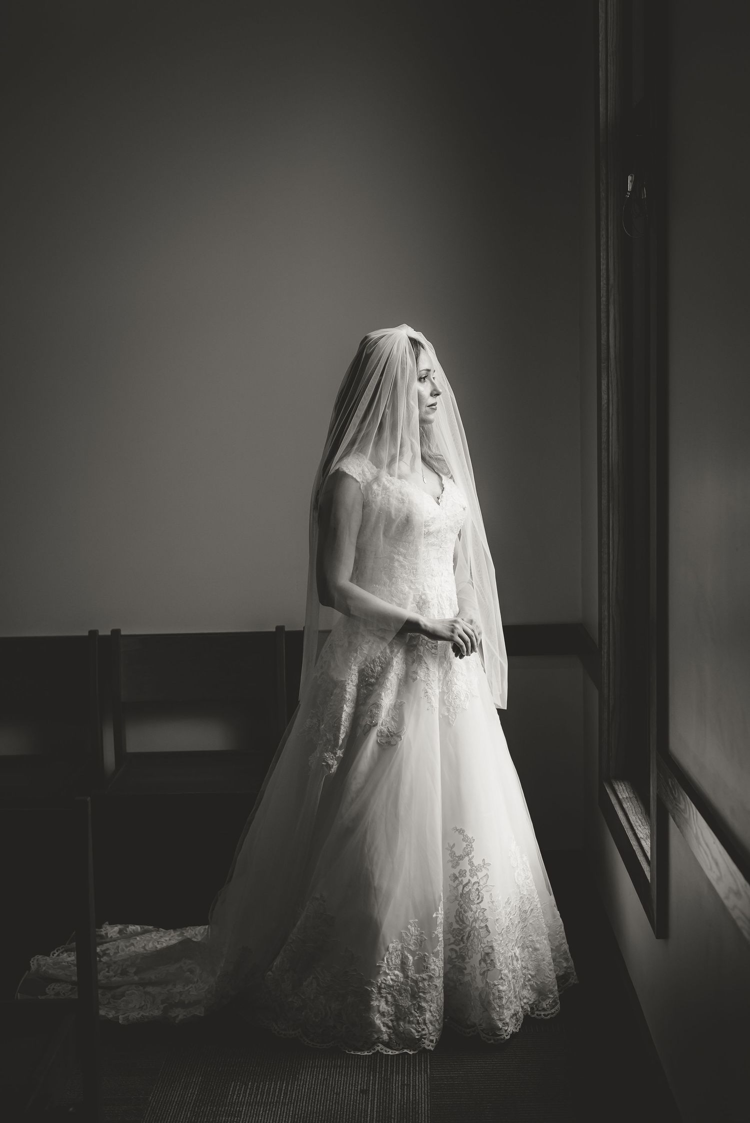 Bride in lace wedding gown and veil stands by window in elegant black and white portrait photo.