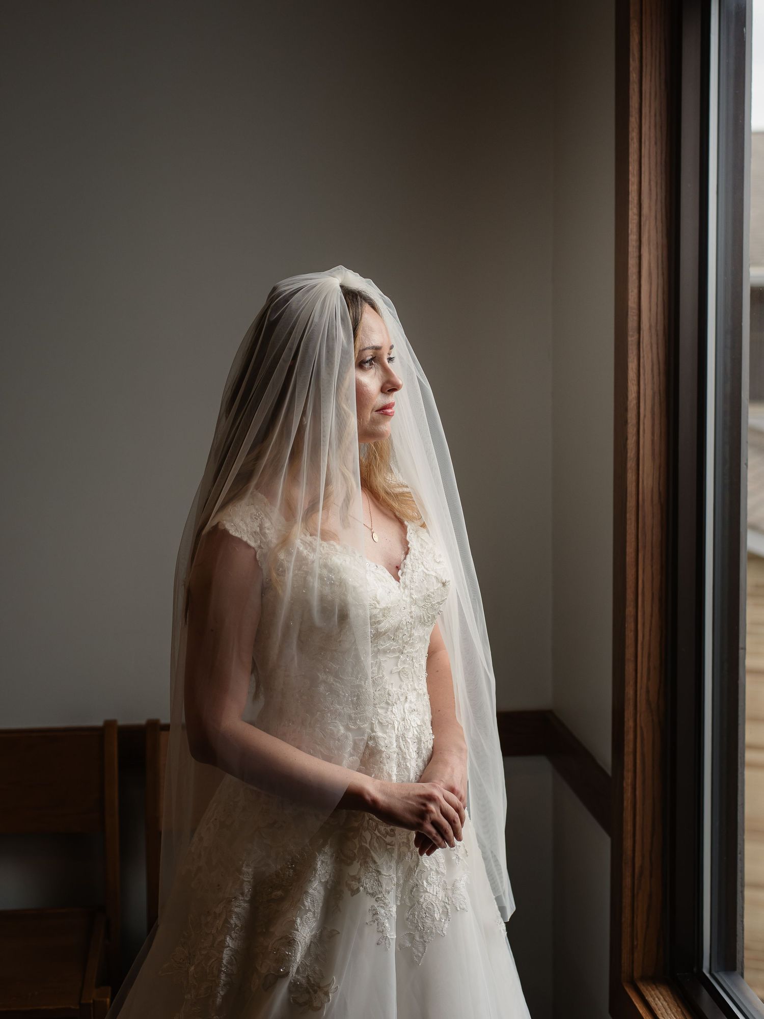 Bride in lace wedding gown and veil gazes out window in soft natural light.