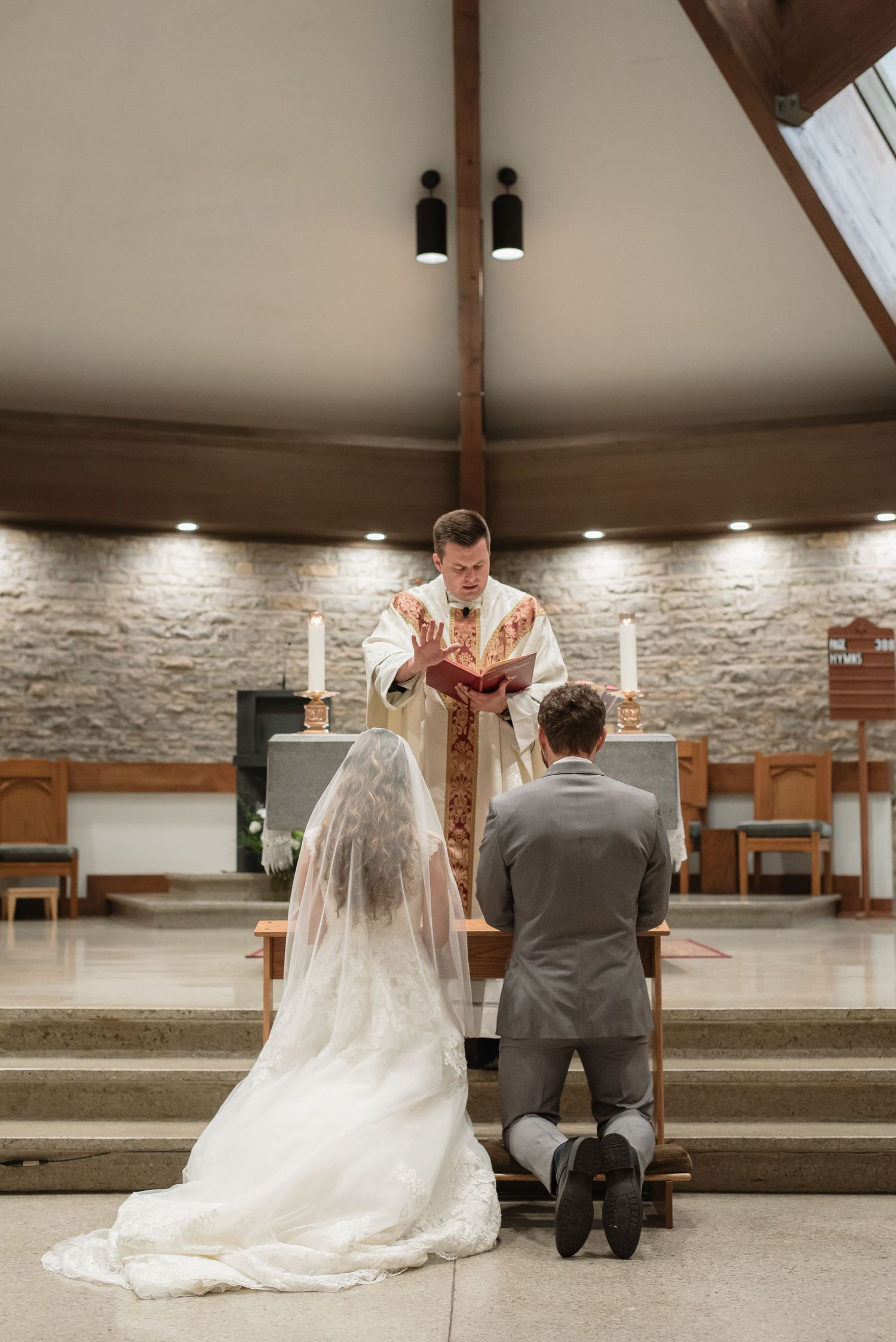 Bride and groom kneeling at church altar during Catholic wedding ceremony with priest offering blessing.
