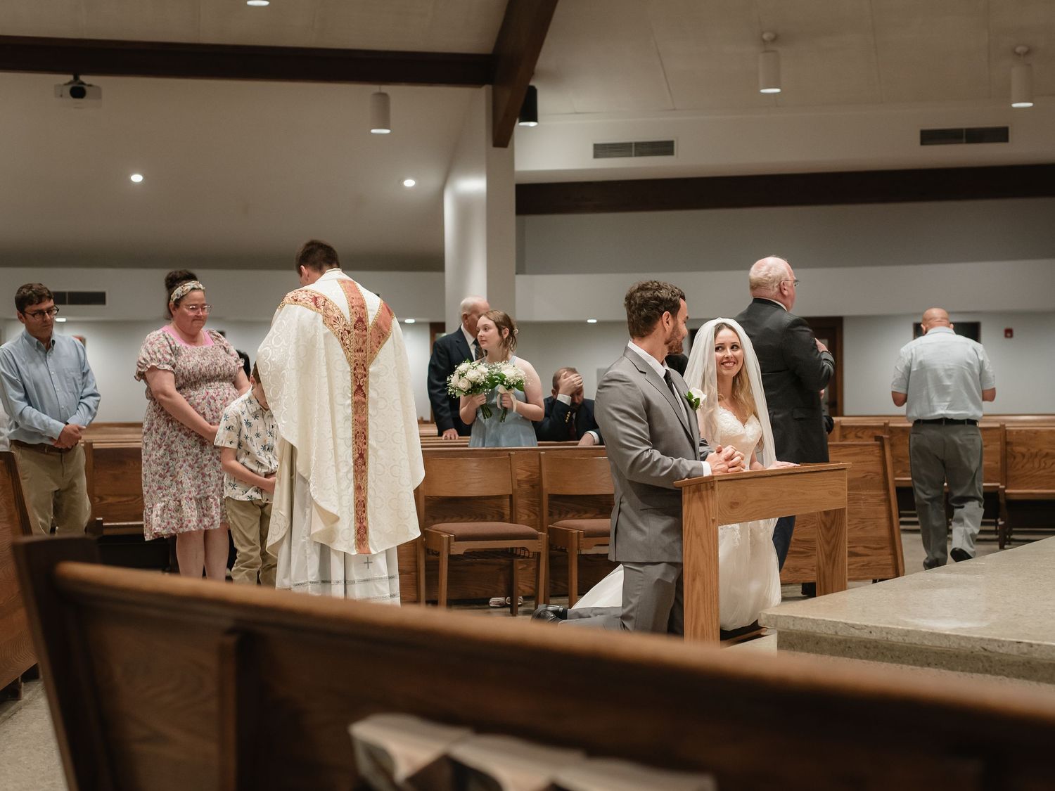 Bride and groom exchange vows at Catholic church wedding ceremony with guests standing in wooden pews.