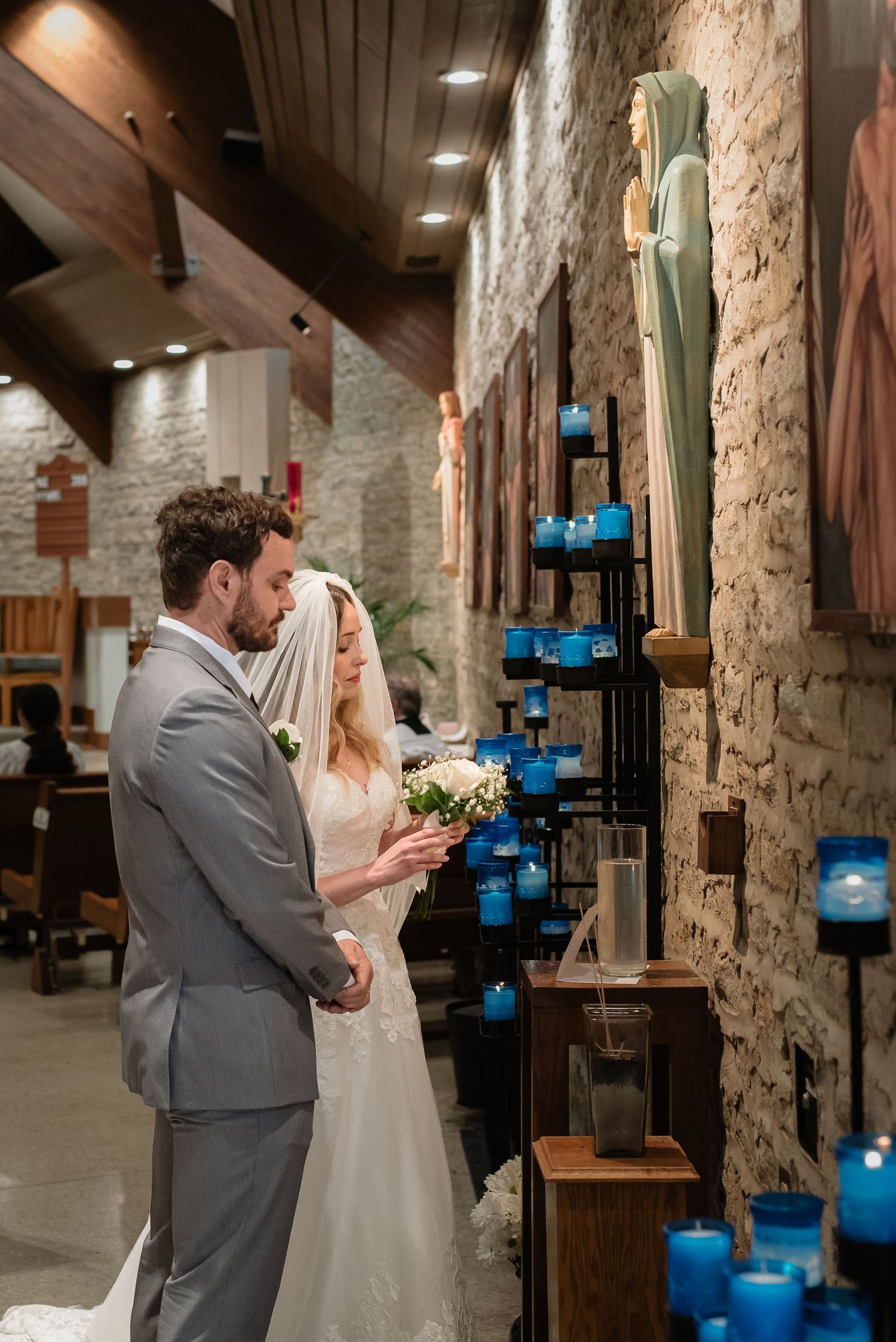 Bride and groom holding hands near a candlelit shrine with a Madonna statue inside a stone church.
