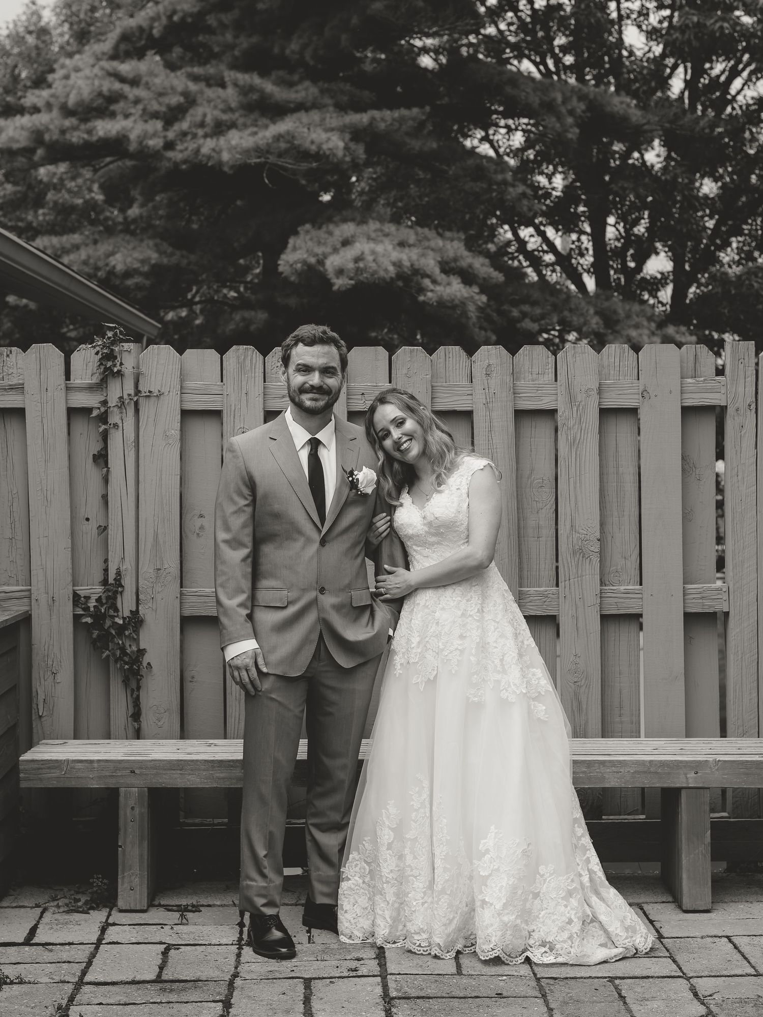 Black and white photo of a bride and groom posing together in front of a wooden fence outdoors on their wedding day.