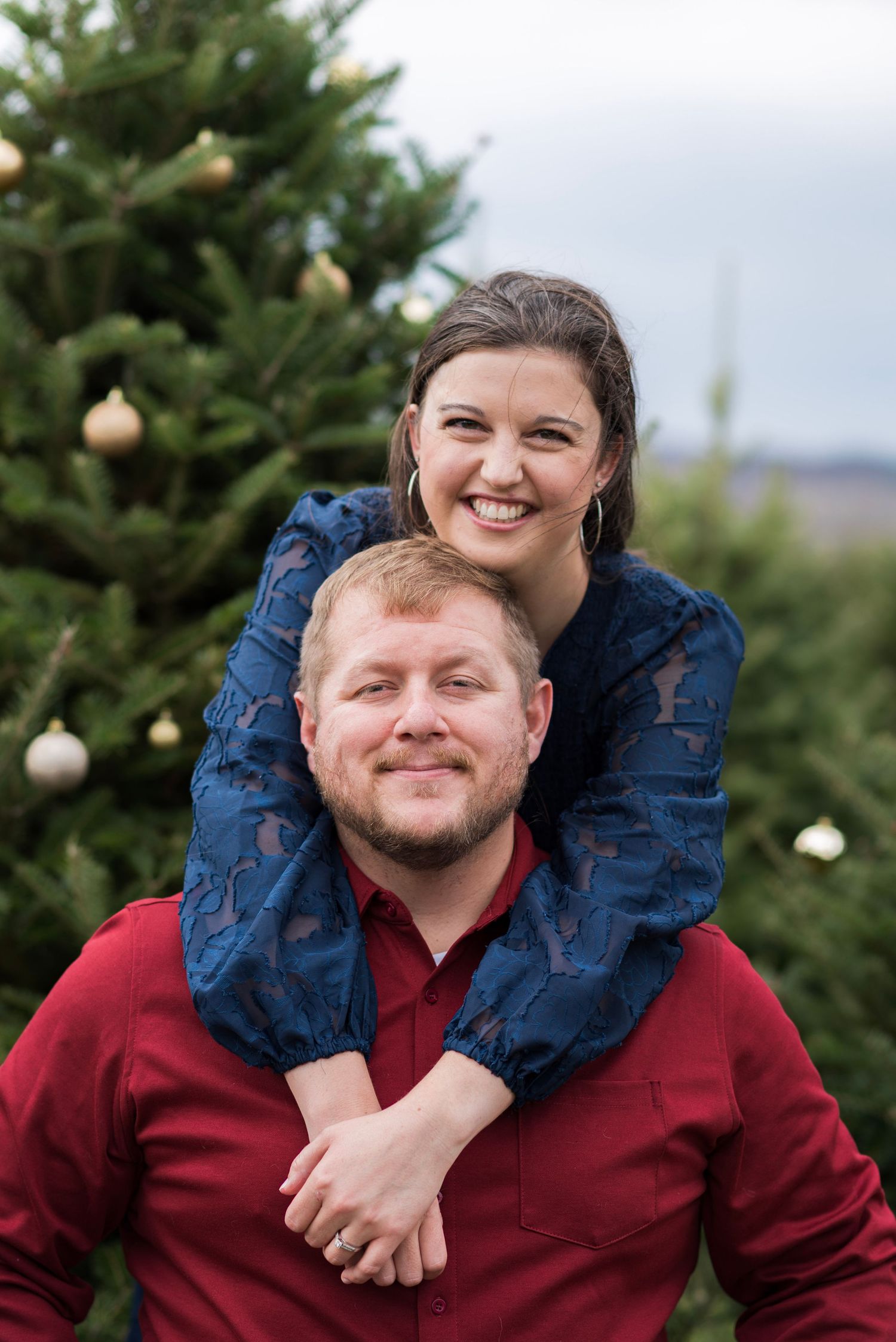 A playful couple portrait at a Christmas tree farm with evergreen trees in the background.