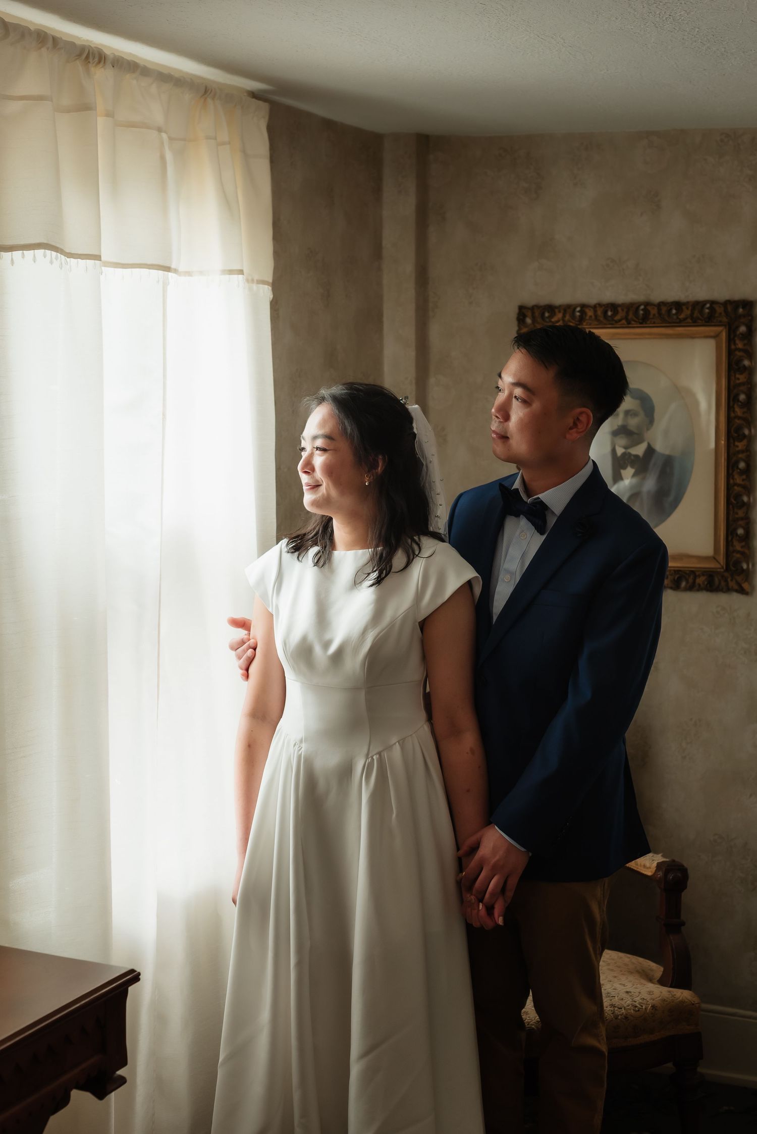 Bride in white dress and groom in navy suit hold hands by a sunlit window in an elegant room.