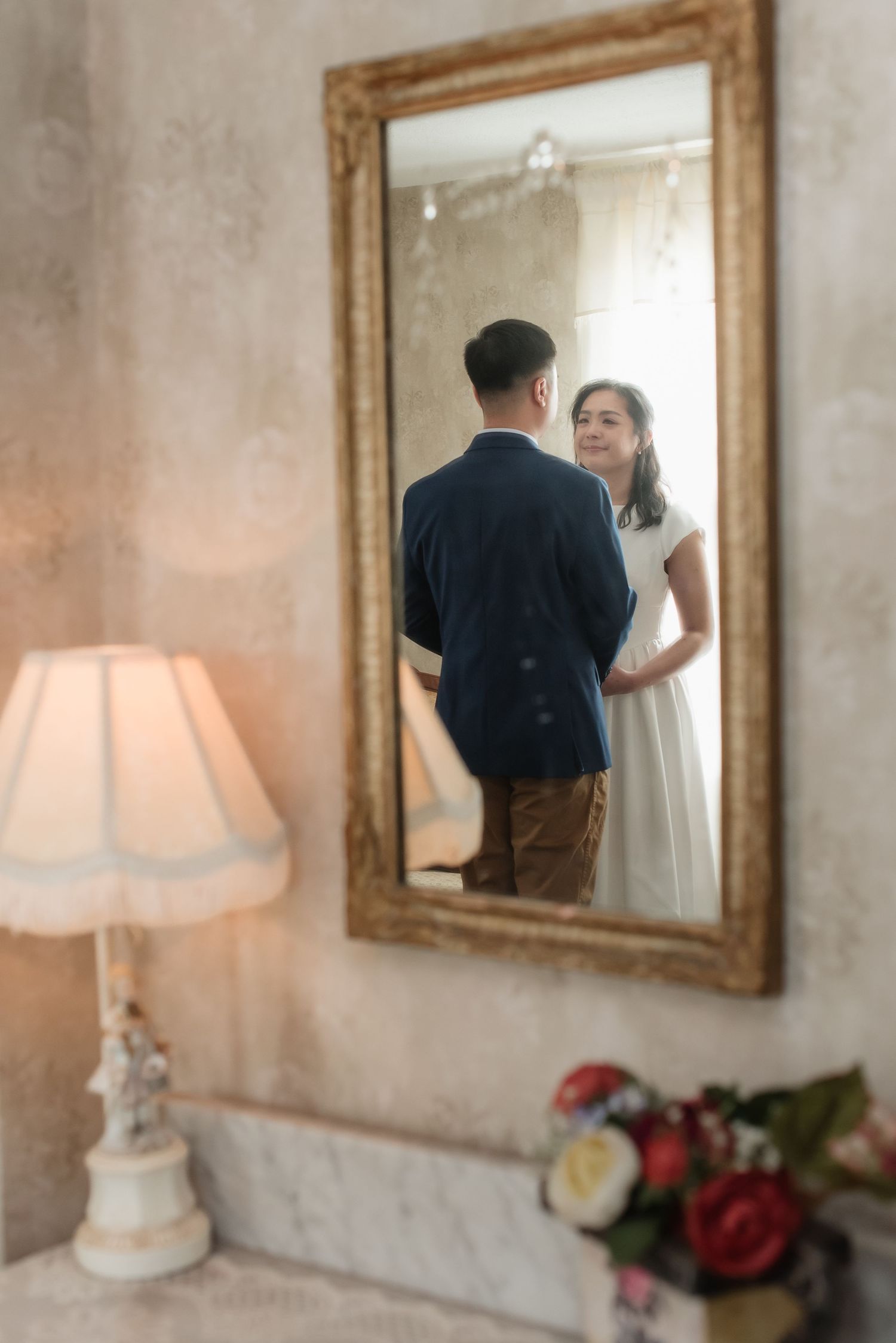 Couple sharing a romantic moment reflected in an ornate gold-framed mirror, with a vintage lamp nearby.