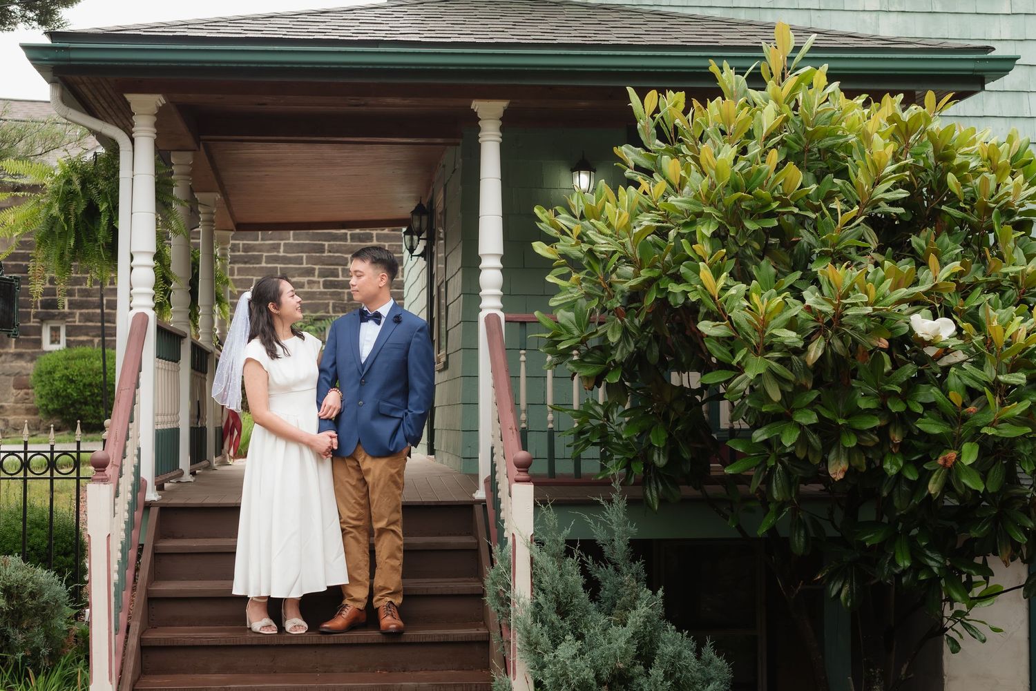 Bride and groom in wedding attire standing on porch steps of a charming home surrounded by lush greenery.