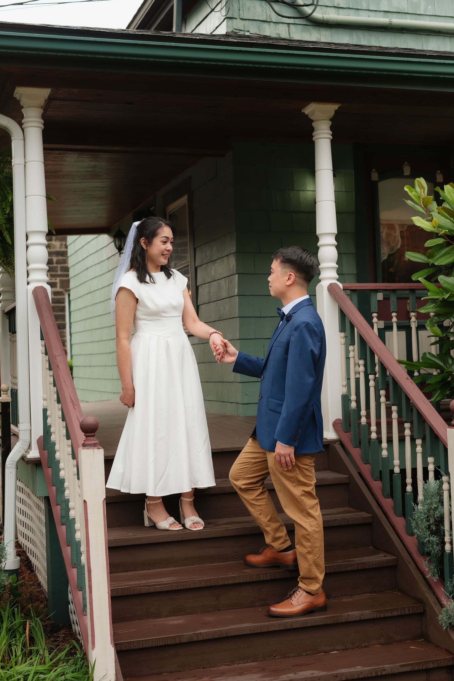Bride in white midi dress and groom in blue suit hold hands on wooden porch steps of a vintage green house.