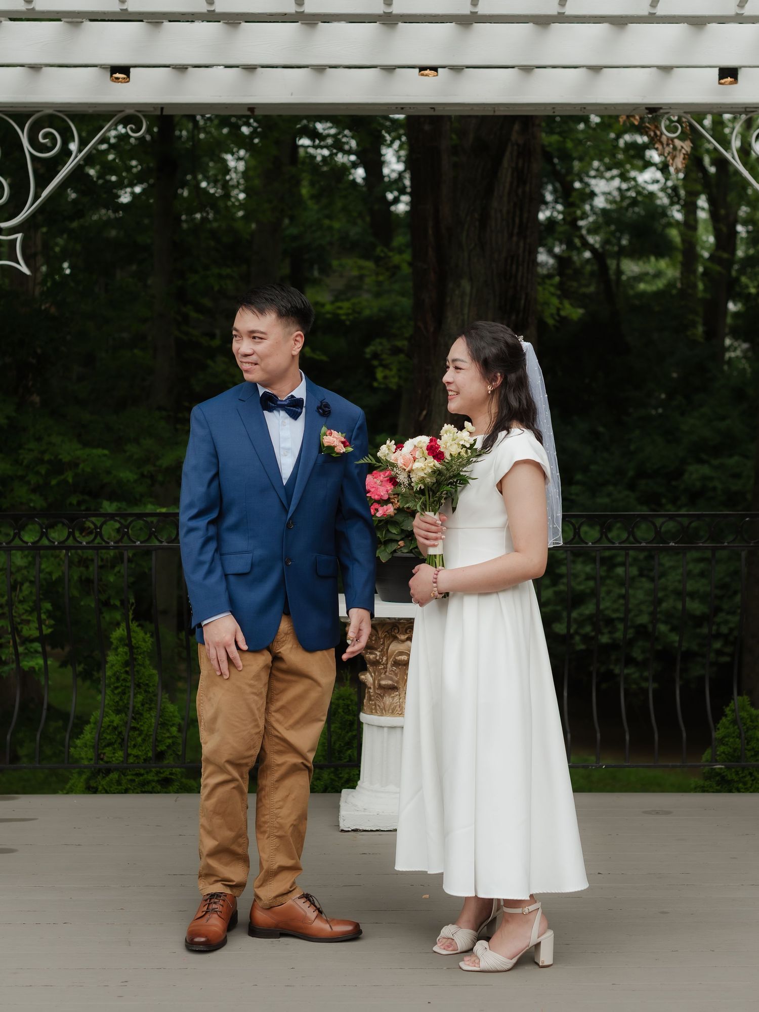 Bride holding colorful bouquet and groom in navy suit stand under garden pergola on wedding day.