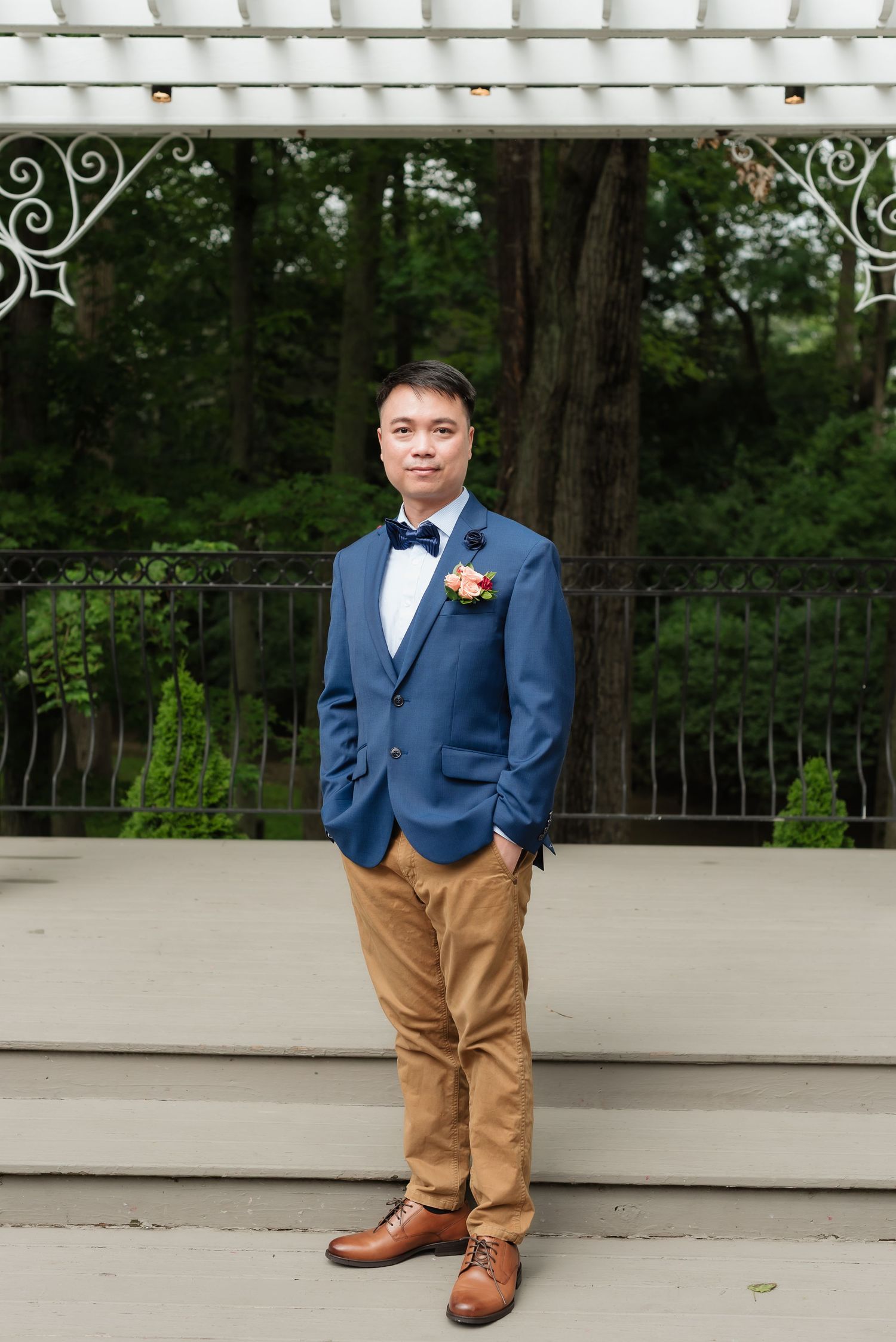 Groom in blue blazer, bow tie, tan pants, and brown dress shoes stands on outdoor venue deck with floral boutonniere.