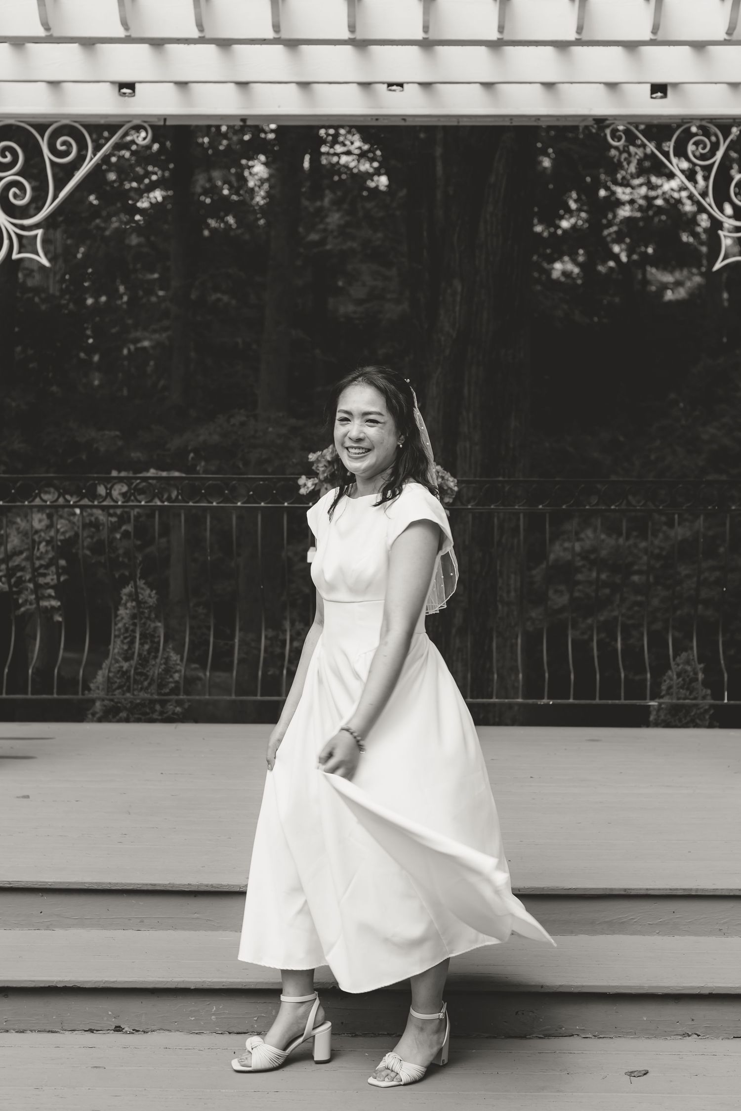 Black and white photo of a smiling woman in a white midi dress and heels posing outdoors near a decorative railing.
