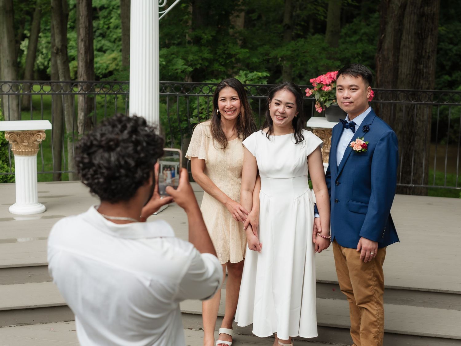 A person photographs three people posing outdoors at a wedding ceremony near white columns and greenery.
