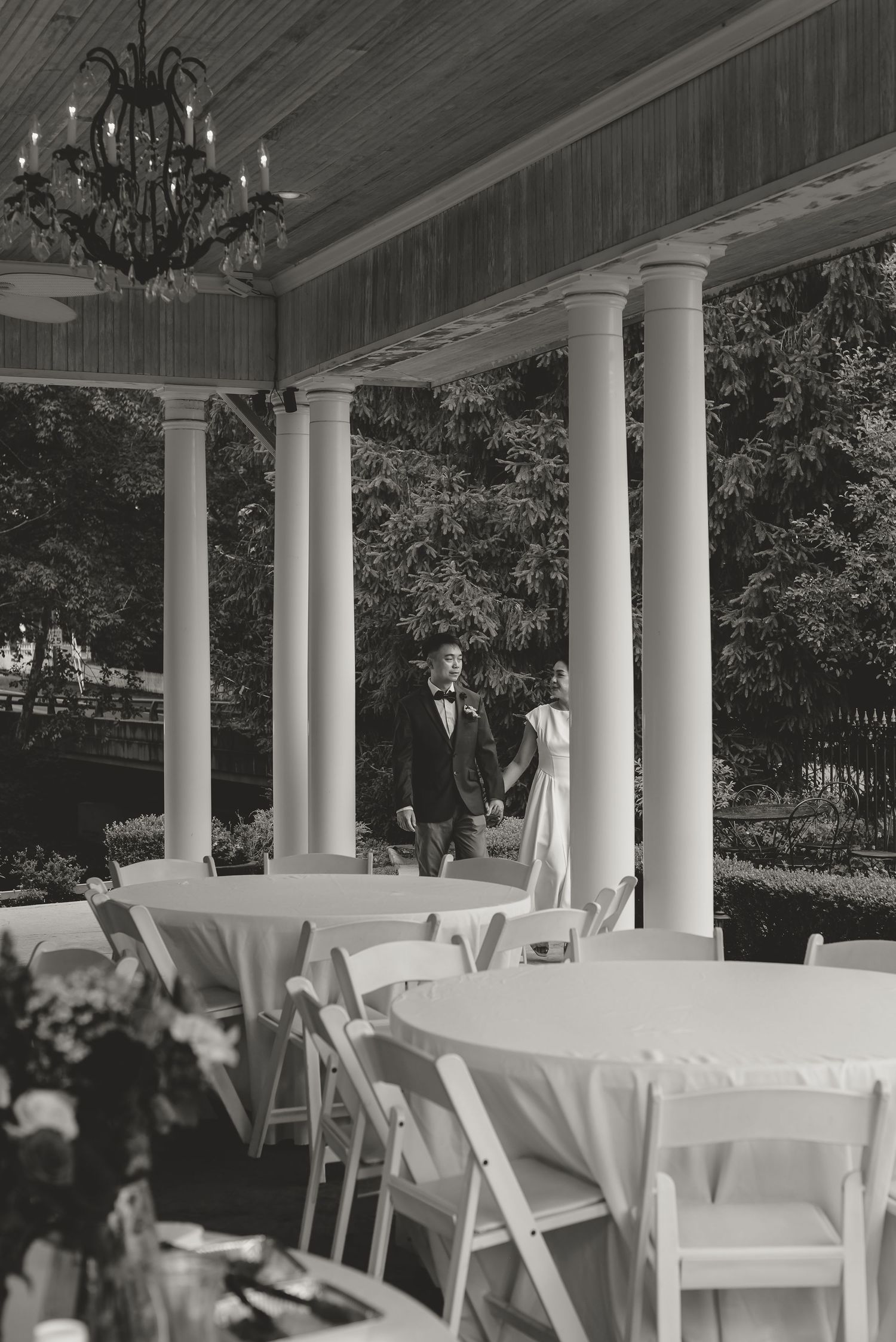 Black and white photo of a couple in wedding attire walking hand in hand on an elegant columned porch with round tables.