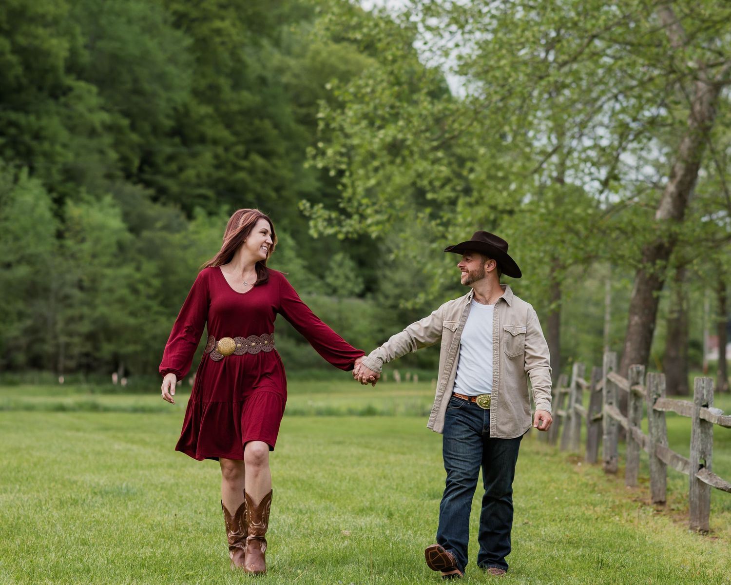 A couple walks together through a lush green park setting with trees and fencing in the background.