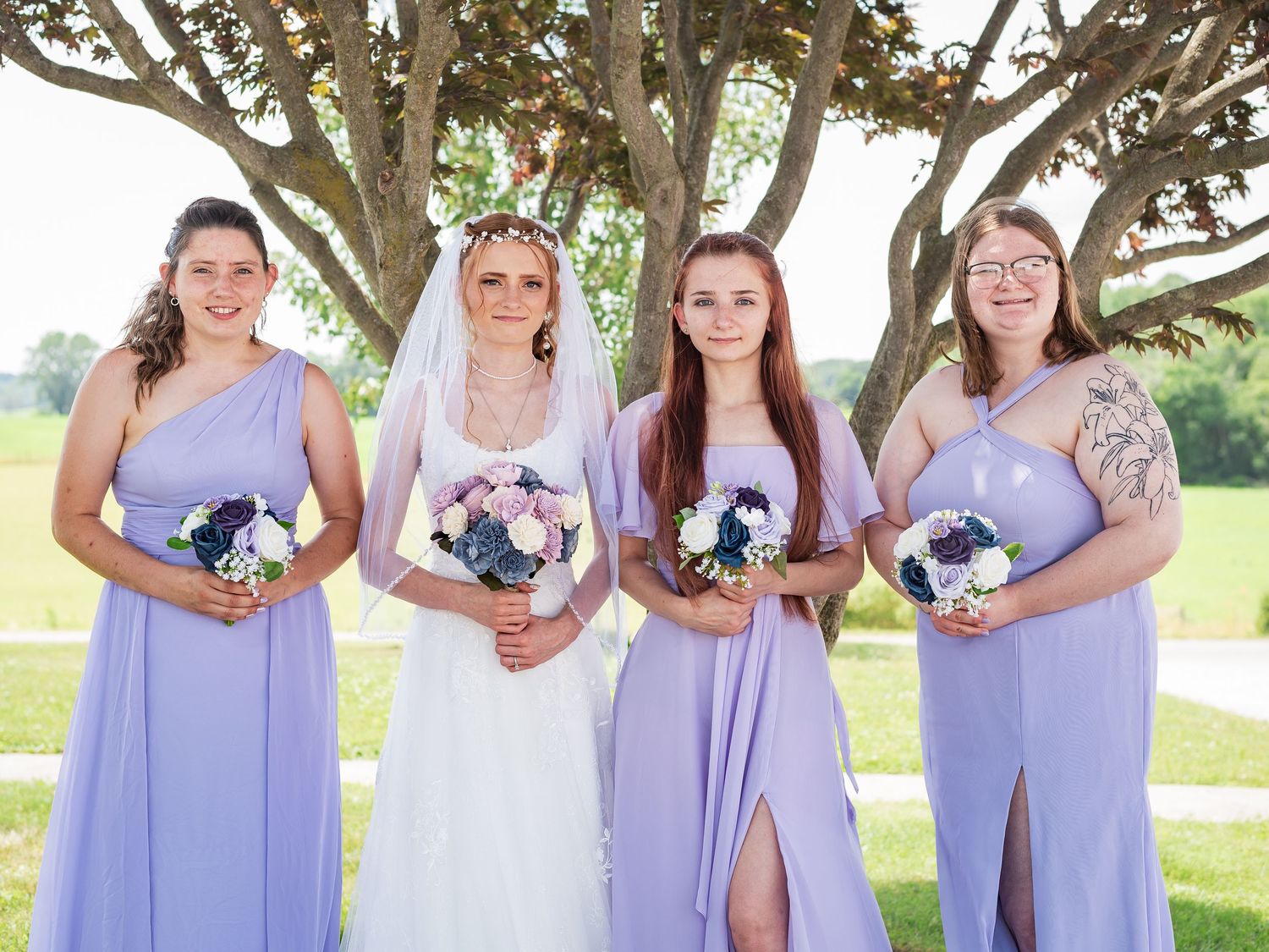 Bridal party in lavender dresses posing with bouquets under a large tree.