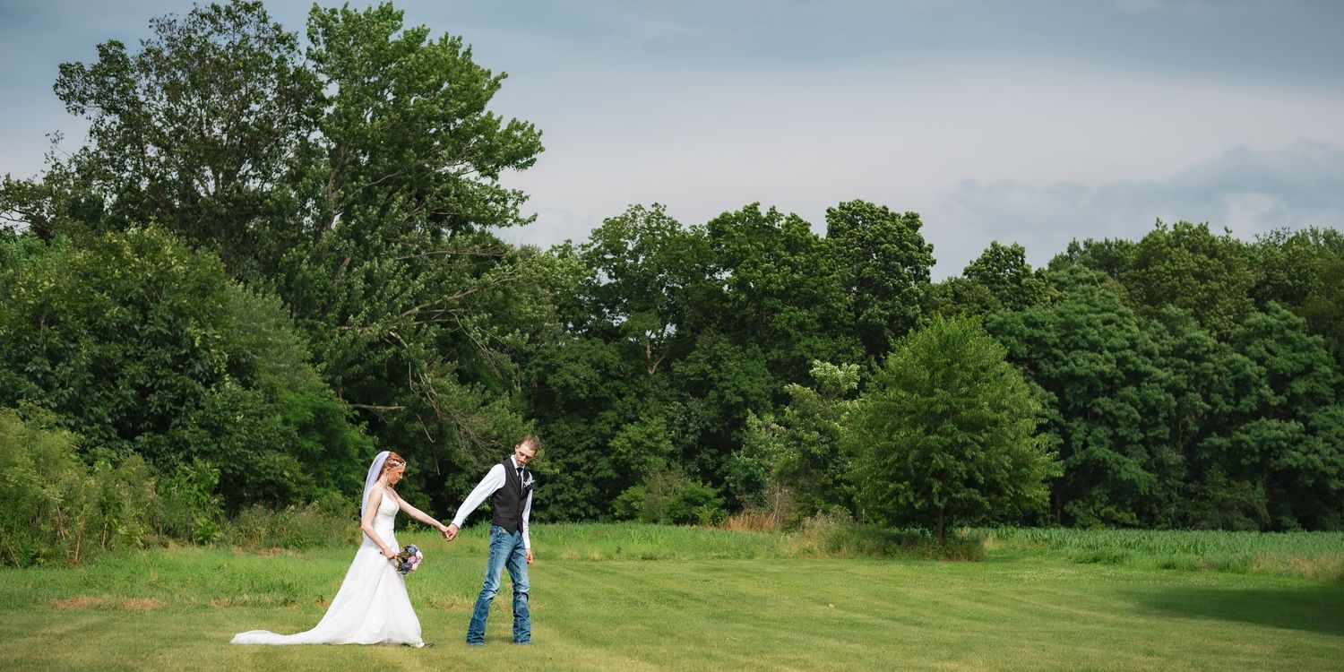 A couple poses in a green field on their wedding day with trees in the background.