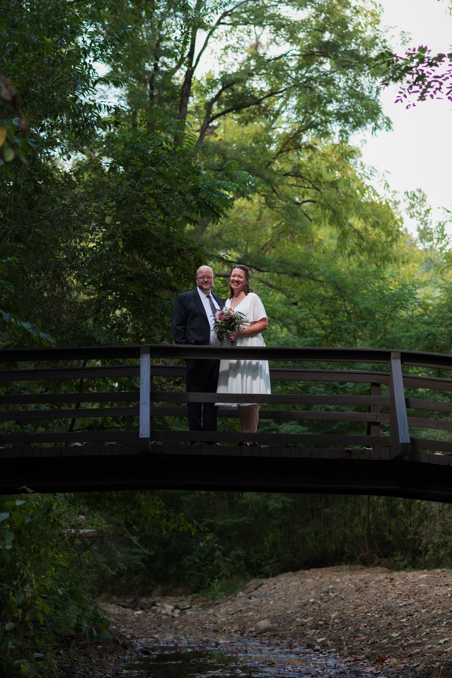 A couple poses on a rustic wooden bridge over a creek surrounded by trees.