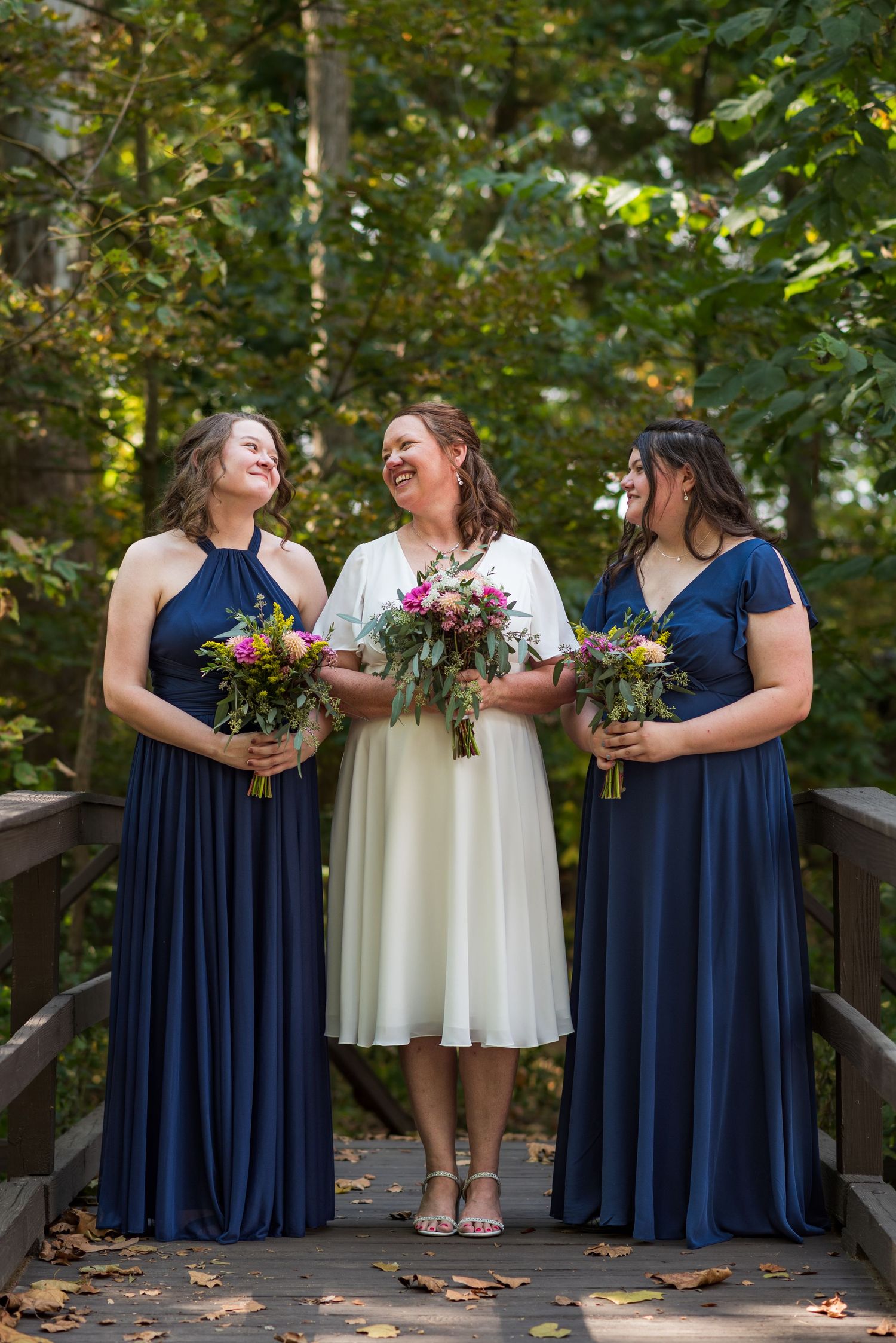 Three women in navy and white dresses share a laugh while holding bouquets on a wooden bridge.