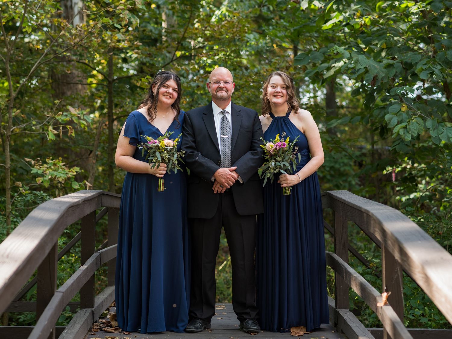 Three people in formal attire pose together on a wooden bridge for a wedding photo.