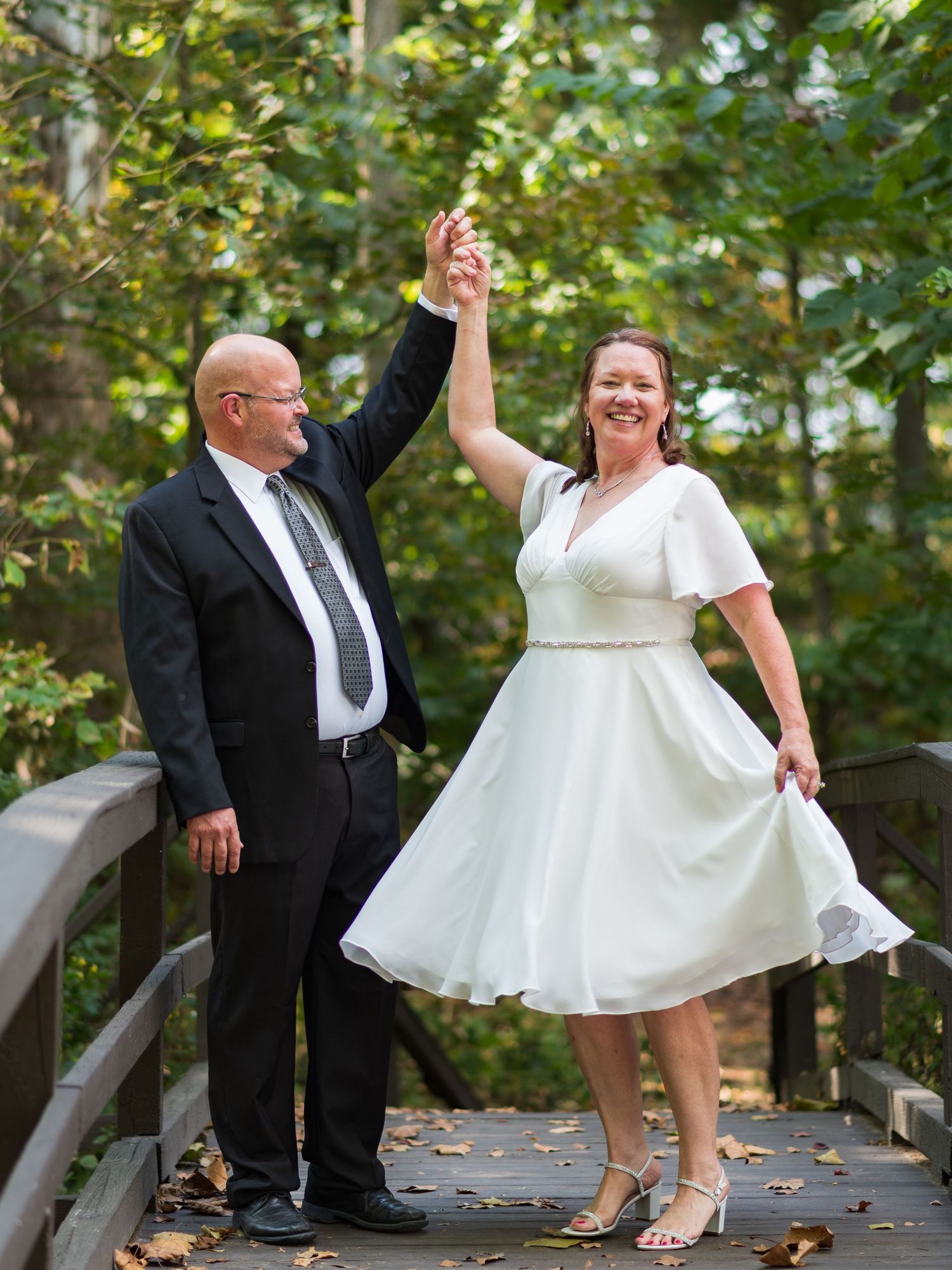 Couple dances joyfully on wooden bridge with autumn leaves scattered around them.