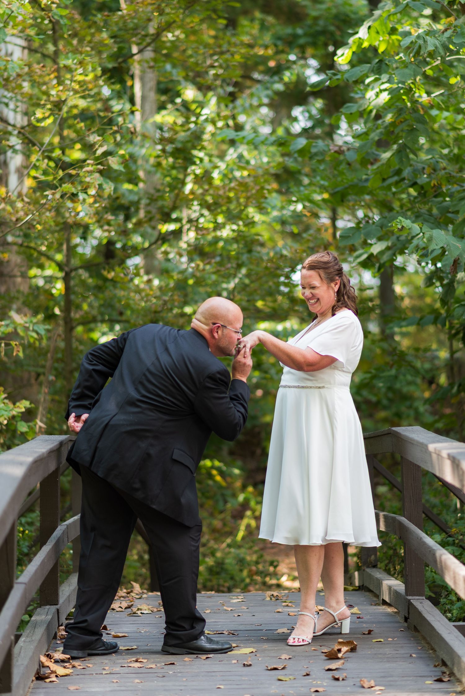 Couple in formal attire shares playful dance moves on a wooden bridge surrounded by autumn foliage.